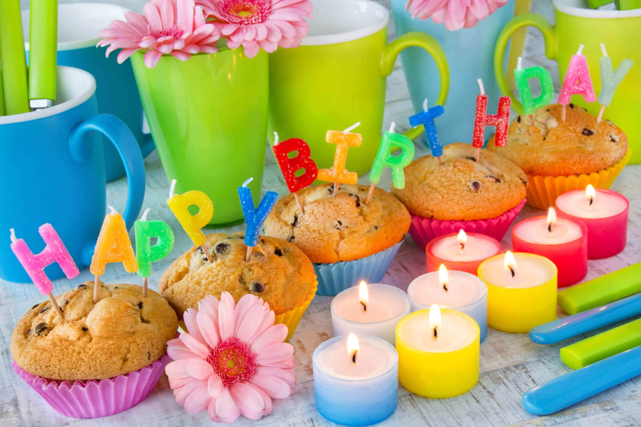 Ein Arrangement aus Tassen (hinten), Muffins mit "Happy Birthday"-Kerzen darin und bunten Teelichtern davor sowie rosa Gerbera-Blumen